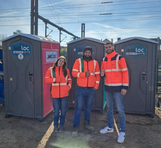 Un homme et une femme en tenue de chantier, fiers, regardent l’appareil photo en levant le pouce devant des toilettes mobiles WC Loc, dont une cabine féminine et une classique.