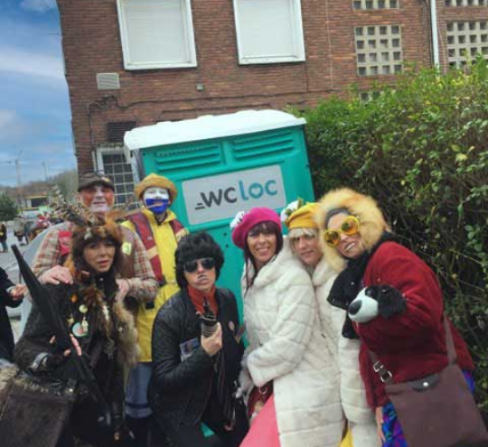 Un groupe de personnes déguisées pose joyeusement devant des toilettes publiques mobiles WC Loc lors d'un carnaval en ville.